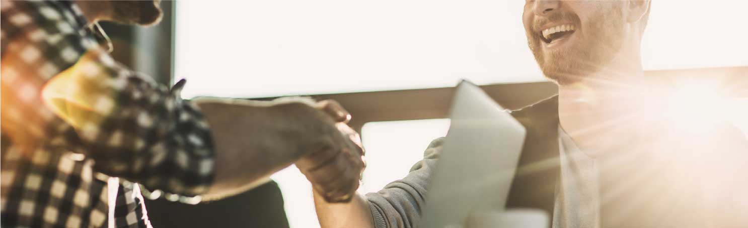 Closeup shot of two men at computers in office smiling and shaking hands.