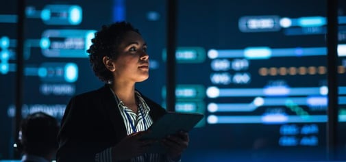A female controller holding a tablet and looking at compliance analytics on large wall-mounted screens.